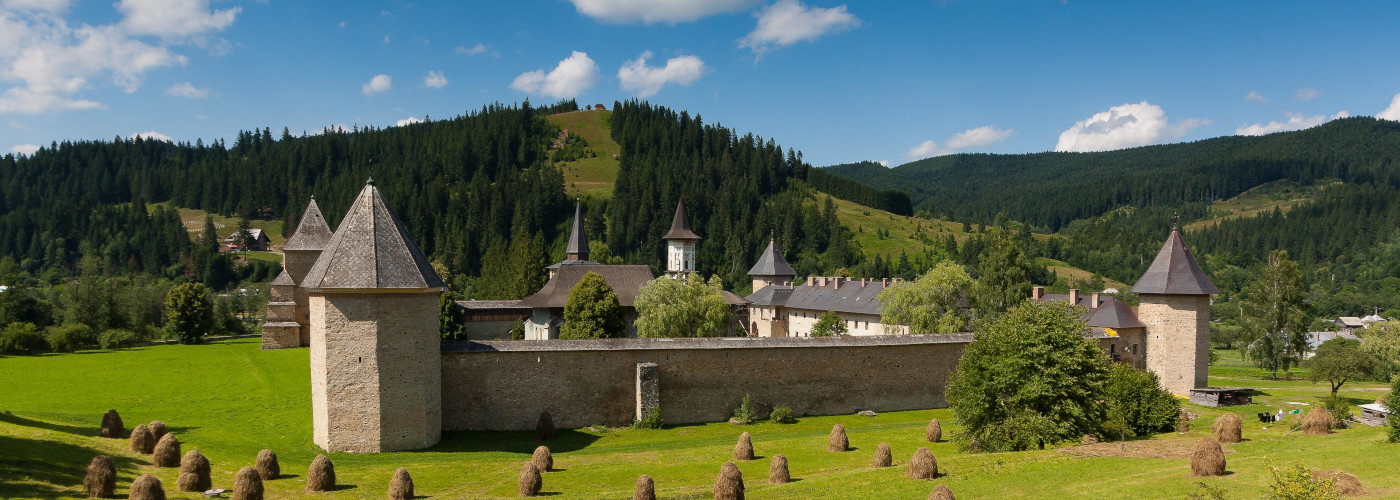 Burganlage mit vier runden Türmen und Wehrmauer vor bewaldeten Hügeln unter blauem Himmel
