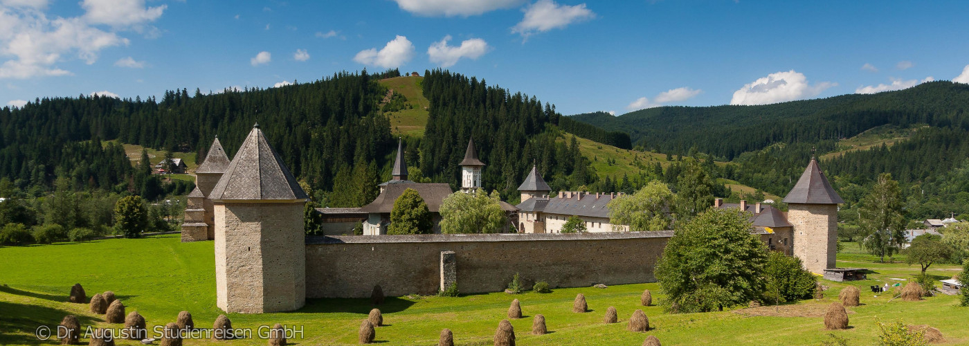 Burganlage mit vier runden Türmen und Wehrmauer vor bewaldeten Hügeln unter blauem Himmel