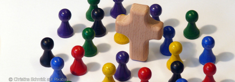 Holzkreuz umgeben von bunten Spielfiguren auf weißer Fläche