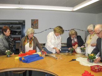 Sechs Frauen stehen um einen großen Holztisch mit buntem Stoff, Scheren und kleinen Topfpflanzen, eine Frau schneidet Folie.