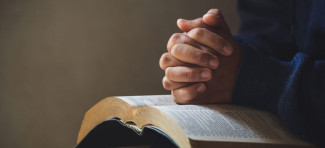 Zwei gefaltete Hände über einem aufgeschlagenen Buch, vermutlich einer Bibel, vor einem unscharfen Hintergrund.