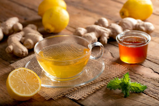 Ein Glas mit gelbem Tee auf Untertasse, umgeben von Zitronenhälfte, Ingwerwurzeln, Honigglas und Minzblatt steht auf einem Holztisch.