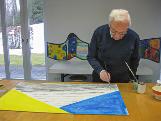 Älterer Mann steht an einem Tisch und malt mit Pinsel auf einer großen Leinwand mit gelbem und blauem Dreieck vor grauen Linien.