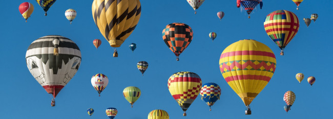 Mehrere bunte Heißluftballons unterschiedlicher Muster schweben vor klarem blauem Himmel.