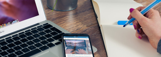 Ein Laptop und ein Smartphone liegen auf einem Holztisch. Eine Hand hält einen blauen Stift und schreibt in ein offenes Notizbuch. Im Hintergrund ist eine Pflanze zu sehen.