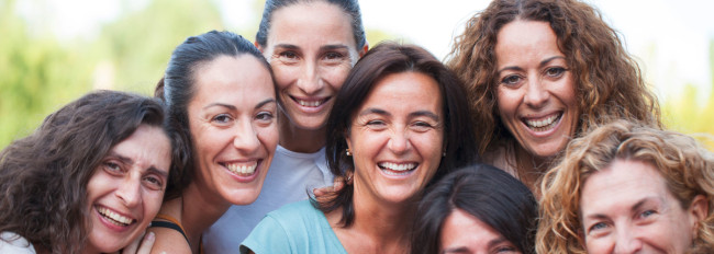 Eine Gruppe von lachenden Frauen in sommerlicher Kleidung vor unscharfem Hintergrund im Freien.