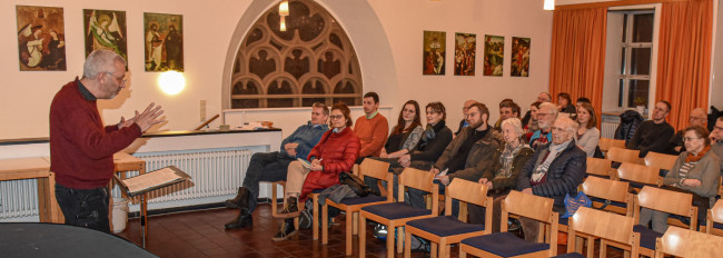 Ein Mann im roten Pullover spricht vor einer Gruppe sitzender Menschen in einem Raum mit Kirchenfenster und religiösen Bildern an der Wand