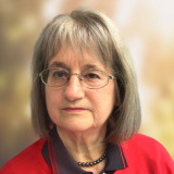 Frau mit schulterlangem grauem Haar, trägt rote Jacke mit schwarzem Kragen und schwarze Perlenkette.