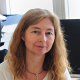 Frau Strunz mit langen, welligen blonden Haaren, trägt eine weiße Bluse und eine Halskette mit mehreren kleinen Anhängern, sitzt vor einem Computerbildschirm und einem Fenster.