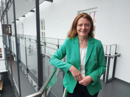 Eine Frau mit grünem Blazer und weißem Oberteil lehnt an einem Glasgeländer in einem modernen Innenraum